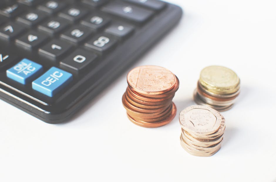 Close-up image of a black calculator with blue and white buttons, situated on a white surface alongside three stacks of coins—one with copper-coloured coins, another with mixed coins, and a third with silver-coloured coins—highlighting financial considerations related to house removals and moving services. The coins are arranged in neat stacks near the calculator, illustrating budget planning or cost analysis for house relocation, with the environment suggesting an indoor setting possibly associated with a professional removal service like Man With a Van Sydenham, indicated by the context of house removals and transparent pricing explanations.