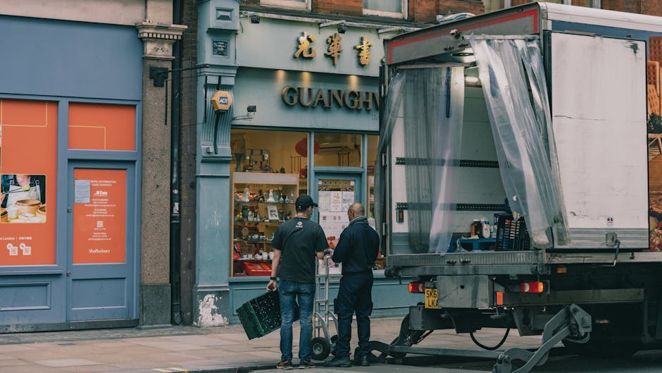 Two men are standing on the pavement outside a storefront with a sign displaying Chinese characters and the word 'GUANG'. One man is dressed in a black shirt and jeans, carrying a black plastic crate,