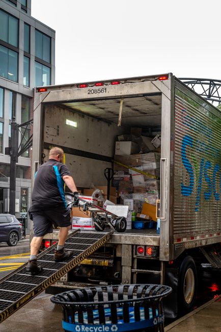 A man wearing a black T-shirt with blue accents and black shorts is loading or unloading a moving truck parked on a city street. The truck's rear doors are open, revealing a variety of packed cardboard boxes, plastic containers, and wrapped furniture inside. The man is pushing a hand trolley with a large rolled-up carpet or rug, preparing to move it into or out of the vehicle. The loading area has a metal ramp with black and yellow safety markings, and a black recycling bin is placed in front of the ramp. In the background, there are modern office buildings with glass facades, a parked vehicle, and the street pavement is wet, suggesting recent rain. The scene captures a professional home relocation or furniture transport process carried out by a moving service like Man With a Van Sydenham.