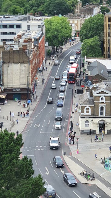 An aerial view of a busy urban street with parked cars and moving vehicles, lined with a mix of residential and commercial buildings, including a red brick storefront and a white building with decorative features. Sidewalks on both sides of the road are populated with pedestrians, some walking and others waiting at crossings. Several bicycles are parked at a bike rack near a corner. Large green trees provide shade along the pavement, and a double-decker bus is visible traveling down the street. The scene depicts a typical city environment suitable for home relocation or furniture transport operations, with visible traffic flow, street furniture, and building facades. This setting would be familiar for a company such as Man With a Van Sydenham conducting house removals or moving services in the area.