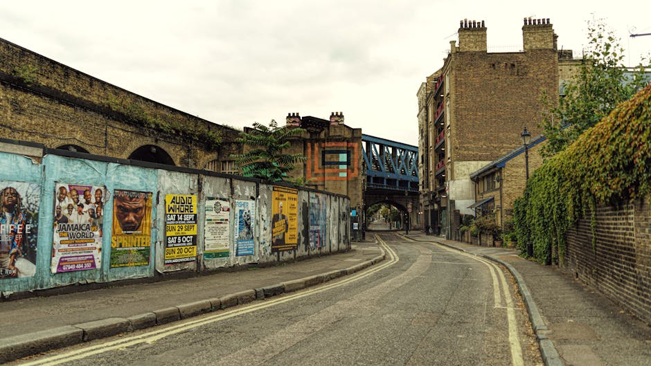 A narrow urban street scene showing a curved asphalt road with double yellow lines, bordered by a sidewalk on both sides. On the left, a weathered plastered wall is covered in multiple colorful posters advertising local events, including music performances and community activities. Behind the wall, part of a brick building with an arched opening and a brick wall extending along the street are visible. To the right, a tall brick wall with lush green foliage growing over it runs alongside the sidewalk. Overhead, a blue metal bridge with latticework spans the street, supported by brick and concrete structures. The scene is captured during daytime with overcast skies, providing diffuse natural lighting. This image depicts an urban environment suitable for home relocations or moving services, where buildings, posters, and infrastructural elements set the context for a typical city street, aligning with the theme of furniture transport and packing and moving processes carried out by Man With a Van Sydenham.