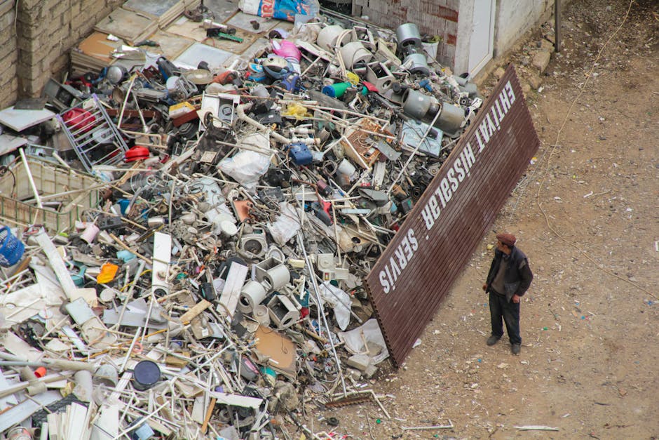 A man standing next to a large pile of assorted household waste and scrap materials, including broken appliances, plastic, metal, and wooden items, situated outdoors on a dirt surface next to a brown metal fence with white text that reads 'THE WASTE MASTER SERVICES.' The waste is contained in a designated area, likely for disposal or recycling, and appears to be part of a large clearing or waste removal process. The man is dressed in dark clothing and a brown hat, observing the debris, which suggests an initial stage of a home relocation or waste collection service. The environment is an open, outdoor space adjacent to a building with stone or brick walls, possibly prepared for collection or disposal as part of a furniture and household items removal project by [COMPANY_NAME]. The scene captures the logistical aspect of clearing bulky waste before loading into a removal vehicle or skip, reflecting typical processes involved in packing, moving, and furniture transport during house removals.
