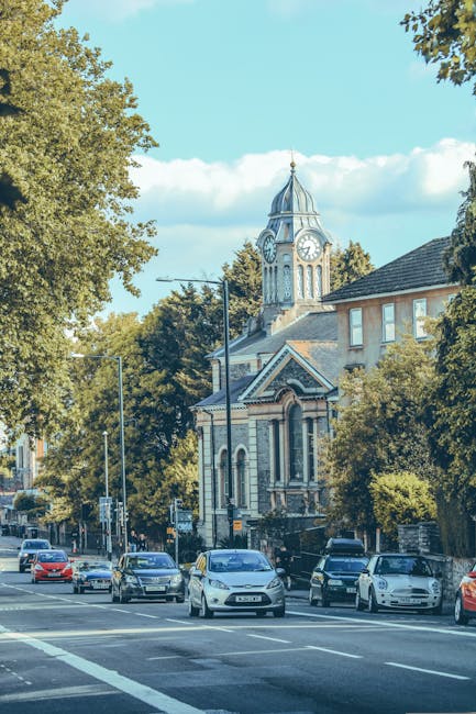 A street scene in Sydenham, Lewisham with parked cars lining the pavement and a church with a clock tower and ornate architecture in the background, surrounded by tall leafy trees under a bright blue sky with some clouds. The trees partially obscure a residential building with large windows and a sloped roof. The image captures the urban environment typical of a neighbourhood undergoing home relocation activities. At the front, the street appears clear, with several vehicles parked along the curb, suggesting possible loading or unloading during a moving process. The presence of a moving company like Man With a Van Sydenham may be associated with the area, offering furniture transport and packing and moving services. The overall scene emphasizes the calm, residential character of Sydenham, with the church serving as a distinctive local landmark amidst the ongoing or planned house removals, vehicle loading, and home relocation logistics, consistent with urban moving operations.