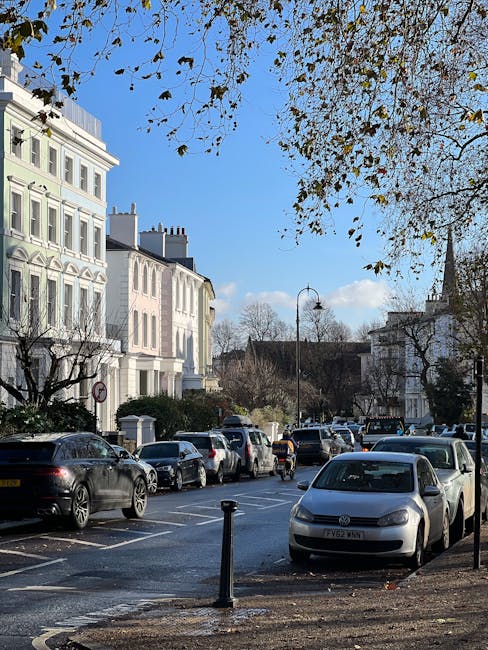 A residential street in Sydenham lined with white Victorian-style terraced houses, with some buildings featuring ornate window frames and decorative facades. The street is filled with parked vehicles, including a silver van positioned close to the curb, which appears to be in the process of being loaded or unloaded for a house removal. In the foreground, there are a few empty parking spaces marked by small black bollards, with some fallen leaves scattered on the pavement, indicating an autumn setting. Overhead, a blue sky with a few scattered clouds provides natural daylight, and a tree with sparse leaves extends its branches across the upper part of the image. This scene captures the typical environment for home relocations and furniture transport services, with an emphasis on the transport process involving the van, which may be affiliated with Man With a Van Sydenham, facilitating smooth and organized moves within the area.
