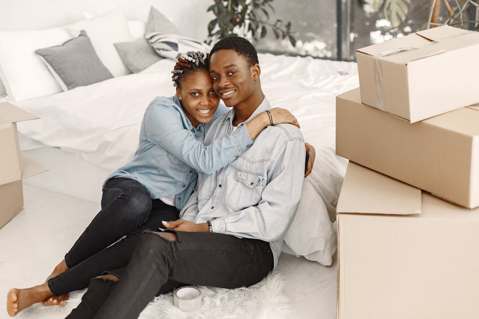A young couple with dark skin, smiling and embracing each other, sitting on a white bed in a well-lit bedroom with white and grey pillows. The woman, wearing a light blue denim jacket and black ripped