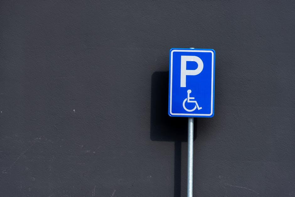 A blue parking sign with a white wheelchair symbol and a capital letter 'P' is mounted on a metal post against a dark grey exterior wall. The sign indicates accessible parking, likely designated for individuals with disabilities. The wall's surface appears smooth and uniform, and there are no other objects or vehicles visible in the image. The sign is positioned in front of the wall, suggesting it is mounted on a paved area such as a driveway or parking space typically used during home relocations. This setting can be associated with moving services provided by Man With a Van Sydenham, especially in contexts requiring parking permits or suspensions for efficient furniture transport, packing, or loading operations during a home move. The lighting is neutral, with subtle shadows cast by the sign, indicating it might be during daylight hours.