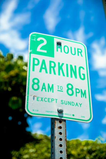 A green and white parking restriction sign mounted on a metal pole, indicating a two-hour parking limit from 8 am to 8 pm, except on Sundays. The sign is outdoors with a background of a partly cloudy blue sky and some out-of-focus green foliage. The sign is part of a street scene related to urban parking regulations. It is relevant for house removals and moving services, such as those offered by Man With a Van Sydenham, as parking permits and restrictions often impact the logistics of furniture transport and home relocation efforts in SE26 areas.
