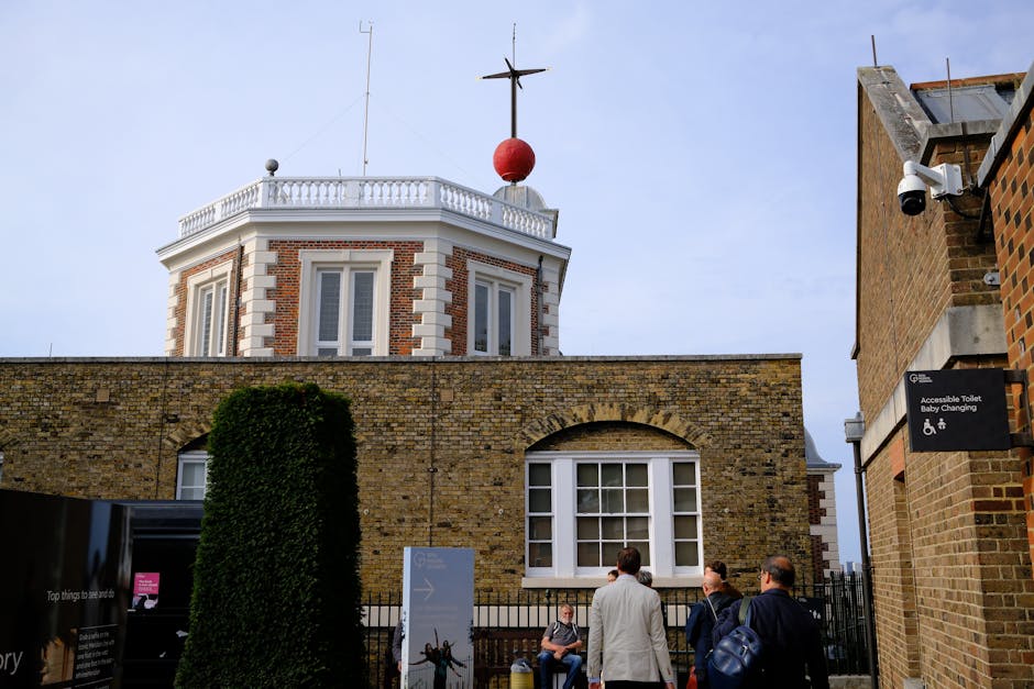 Photograph of a group of four people standing outside a historic brick and white stone building with a rounded tower topped by a weather vane and red ball. The building features large white-framed windows and is surrounded by a low brick wall, with a neatly trimmed bush visible in the foreground. A security camera is mounted on the wall of a neighboring building on the right, which also displays a sign indicating accessible toilet and baby-changing facilities. The scene suggests a setting for house removals or a furniture transport process, possibly part of a client consultation or planning stage for a local home relocation near Horniman Museum. The individuals are dressed in business casual attire, engaged in conversation, and appear to be in an outdoor area associated with a professional moving service such as Man With a Van Sydenham, involved in packing and moving logistics.