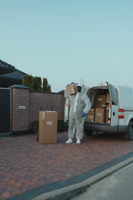 A white commercial van with a closed cargo area is parked on a narrow urban street, with its front facing the camera. The van is positioned close to a peach-colored building with large windows on the 
