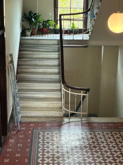 A staircase inside a residential property with a marble-tiled landing and wooden stairs leading up to an open doorway. The staircase features a dark wooden handrail and white balustrades, with natural light coming through a window at the top. On the left side, a silver step ladder leans against the wall, and several potted plants are positioned on the window sill, adding greenery to the space. The wall beside the stairs is painted in a neutral light colour, and a spherical ceiling light fixture is visible on the right. This scene depicts interior elements of a home in the context of a house move, with potential focus on the challenges of transporting furniture and belongings through the staircase during a relocation managed by Man With a Van Sydenham.