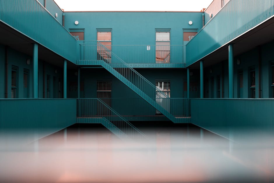 View of an outdoor internal stairwell within a modern, multi-storey residential building. The structure features teal-colored metal railings and staircase, with several doorways on each floor, some open and some closed. The staircase zigzags between floors, providing access to different levels, and the overall environment appears clean and well-lit, highlighting the building's architectural design. The image captures the inside of a home relocation setting where furniture or boxes might be transported between floors. Man With a Van Sydenham specializes in removals and furniture transport services, including navigating complex staircases like this during house moves and packing and moving processes.
